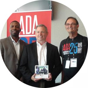 A group of three men stand in front of a roll-up ADA25 banner. The man in the middle is holding an award with a photo on it. All three men are looking towards the camera posed.