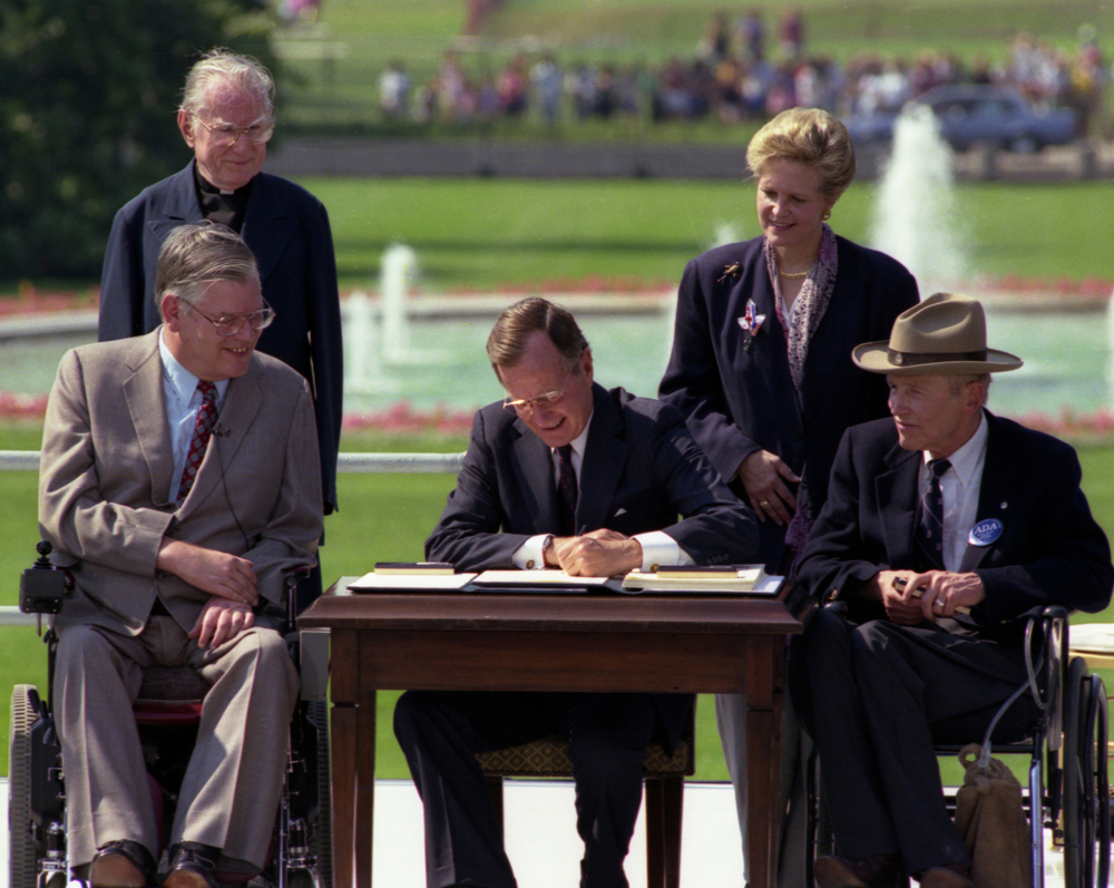 President Bush signs the Americans with Disabilities Act on the South Lawn of the White House. Sharing the dais with the President and he signs the Act are (standing left to right): Rev. Harold Wilkie of Clairmont, California; Sandra Parrino, National Council on Disability; (seated left to right): Evan Kemp, Chairman, Equal Opportunity Commission; and Justin Dart, Presidential Commission on Employment of People with Disabilities