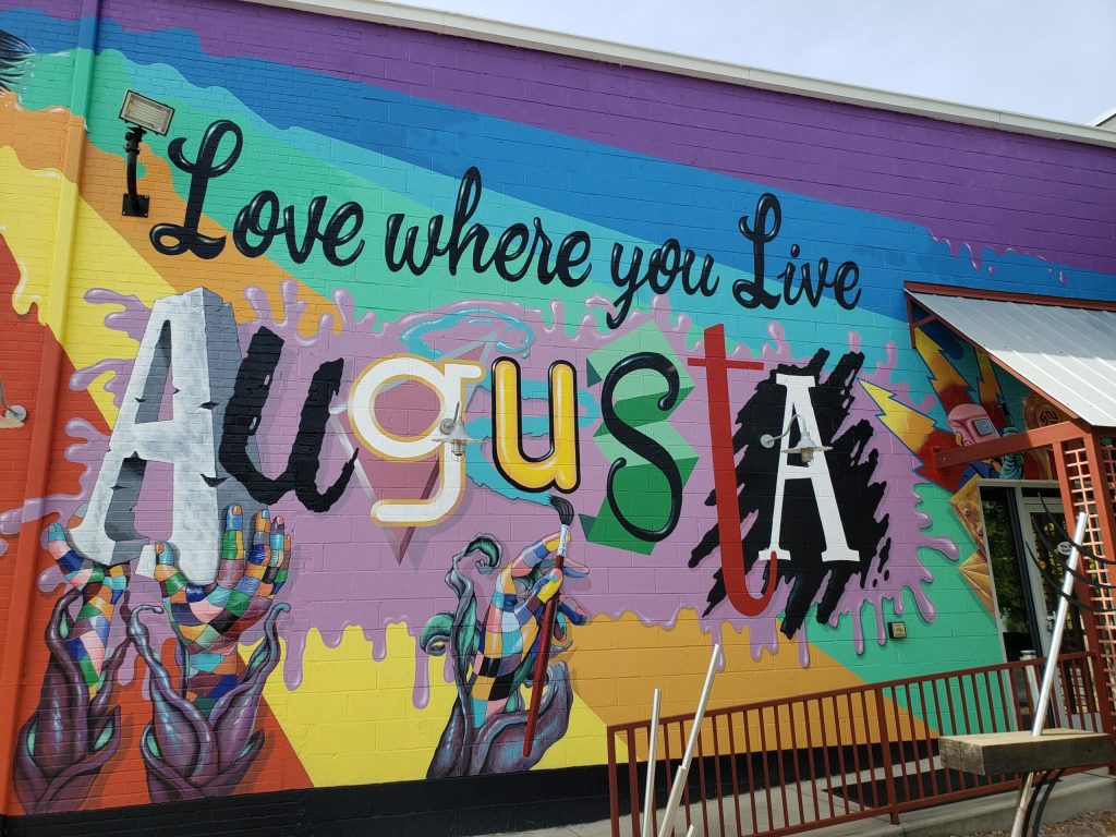 The image is of a mural on the side of 523 Gallery in Augusta, GA. It says "Love Where You Live Augusta." The background has rainbow colors with hands sculpting and painting.