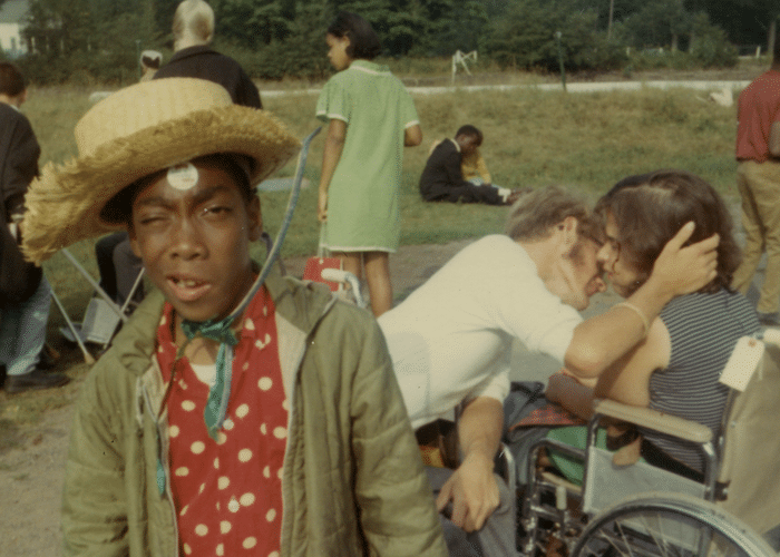 Young boy with a straw hat squinting  at the camera. Behind him, a boy and girl, both in wheelchairs, embrace.