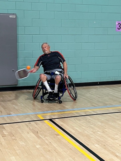 Photo of man in a wheelchair swinging to hit a pickleball. The background is a cinderblock wall painted teal.