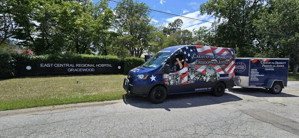 The Caravan for Disability Freedom and Justice. A woman sits in the driver's seat with her arms crossed in an X with the East Central Regional Hospital - Gracewood entry sign in back.