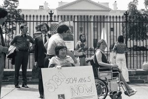 Black and white image of people protesting outside of the white house. A woman (Janet Huemann)  holding a sign "sign 504 reg. s now"